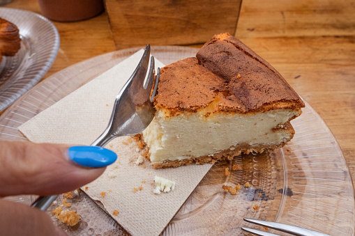 Breakfast. Woman's hand with fork takes a piece of cheesecake on a glass plate, on the table there is a cup of coffee on a plate with metal spoon, glass plate with a croissant and cup of tea. All on a natural wood table. Gastronomy. Tenerife, Canary Islands, Spain
