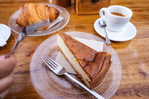 Breakfast. Woman's hand with fork is going to poke cheesecake on a glass plate with a knife and fork, cup of coffee on a plate with metal spoon, glass plate with a croissant and cup of tea. All on a natural wood table. Gastronomy. Tenerife, Canary Islands, Spain