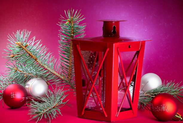A close-up photograph of a red Christmas lantern set next to of spruce branches and colored baubles.