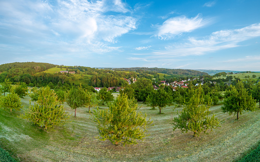 Panorama on valley with green endless fresh meadows, agricultural crop fields, dense mixed wild forest and lonely farms against a cloudy blue sky in the evening sunlight