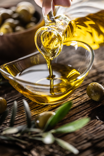 Pouring olive oil from carafe to the glass bowl and in background are olive branches put on table.