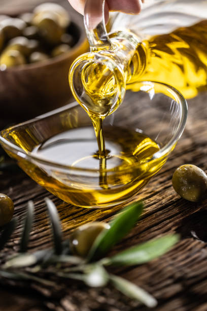 оливковое масло наливаем из графина в стеклянную чашу, а на заднем плане оливковые ветви, поставленные на стол. - cooking oil oil pouring olive oil стоковые фото и изображения