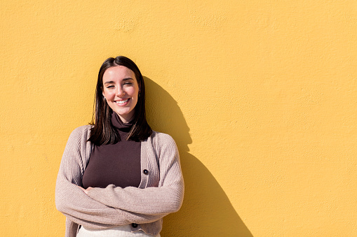 young woman smiling happy looking at camera with arms crossed leaning in a yellow background, concept of youth and lifestyle, copy space for text
