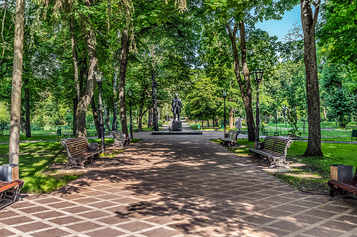 Trostyanets, Sumy Oblast, Ukraine - June 18, 2023: Alley and Monument to Tchaikovsky in the Trostyanets central park. Benches along the footpath among the summer greenery in the city garden