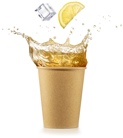 Flying lemon slice and ice cube falling into splashing iced tea in disposable paper cup. Refreshing beverage in eco friendly to go packaging. Real shot.