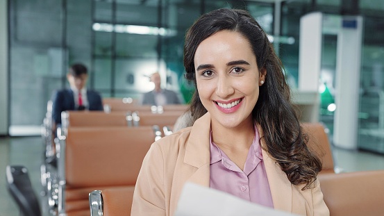 Close up smiling face of young passenger hispanic latin women sitting in terminal gate at airport while waitting airplane flight looking at camera. Travel concept