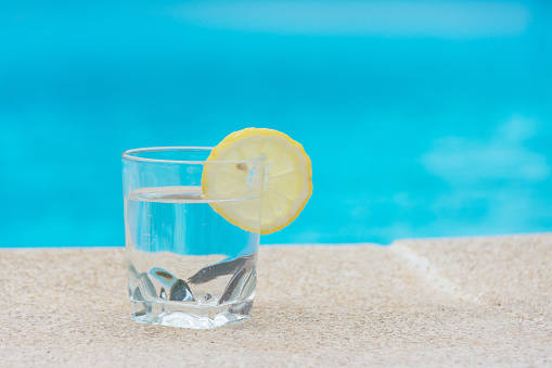 Quench your thirst in the summer to beat the heat. A glass of water with a slice of lemon on the edge of a swimming pool