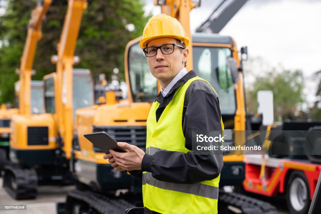 Engineer in a helmet stands next to construction excavators Engineer in a helmet with a digital tablet stands next to construction excavators Fleet of Vehicles Stock Photo