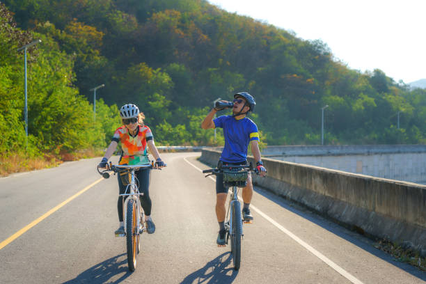 Tired Asian cyclist man drink water from a bottle while riding together for exercise around the lake in the morning