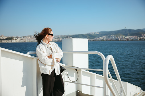 Young tourist woman moving around Turkey, using a ferry boat. She is wearing sunglasses, watching the Izmir panorama around her.
