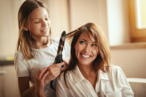 Little daughter is having fun playing hairdressing salon with her young mother, curling her hair with electric hair curler.