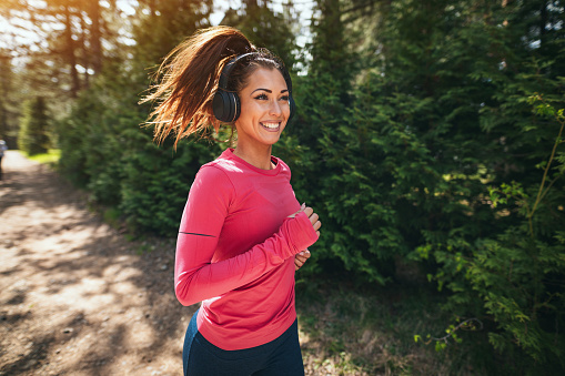 Young beautiful happy female runner listening to music while jogging along a sunny trail in forest.