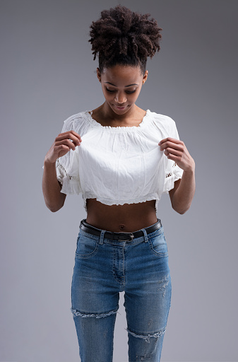 Did I stain myself? Young woman examines her shirt for cleanliness, looking down, hands on dress. Beautiful dark-skinned girl with afro hair, wearing blue jeans and a white crop top, revealing a flat