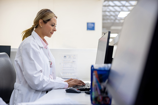 Latin American female doctor using a computer at the reception desk while working at the hospital