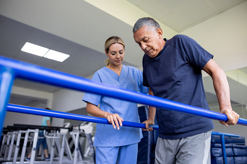 Latin American man doing physical therapy and walking on bars with the assistance of his physical therapist