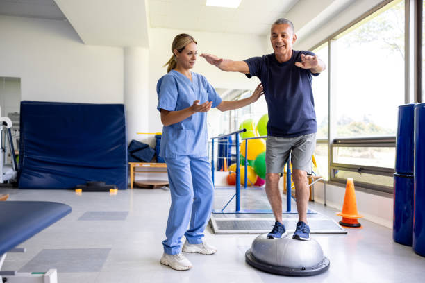 Man doing physical therapy exercises using a balance ball Mature man doing physical therapy exercises using a balance ball with the guidance of his therapist standing had to knee pose stock pictures, royalty-free photos & images