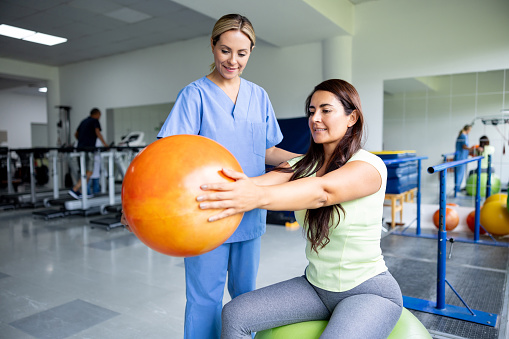 Latin American woman doing physical therapy exercises using a fitness ball with the guidance of her therapist