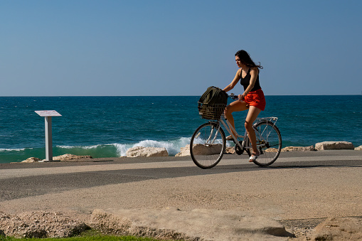 Tel Aviv, Israel - September 15th, 2022: A young woman riding her bicycle on the Tel aviv promenade by the sea, on a summer day.