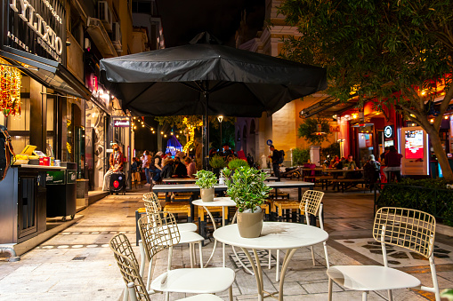 Illuminated sidewalk cafes and shops at night in the lively Monastiraki district near Syntagma Square in Athens Greece. An empty table and chairs with a small plant in focus in the foreground. Lively Monastiraki is known for iconic landmarks including the ruins of Hadrian’s Library, the Ancient Agora and the rebuilt Stoa of Attalos, with a museum exhibiting Athenian artifacts. Monastiraki Flea Market is a jumble of shops selling artisanal soaps, handmade sandals and souvenir T-shirts. The surrounding streets are crammed with traditional tavernas and restaurants, many with Acropolis views.