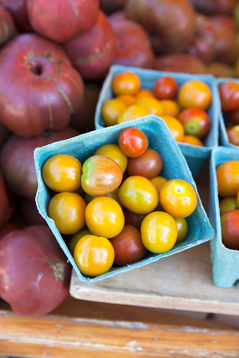 Cartons of freshly-picked yellow and red cherry tomatoes with red heirloom tomatoes in the background at a farmer's market.