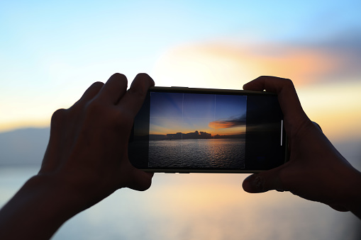 Close-up of a woman's hands with a phone taking photos of a bright sunset and the sea.Hand holding a smartphone taking a photograph of the sea at sunset