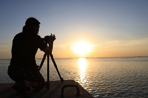 silhouettes of a woman taking pictures against the sunsets. photographer taking a sunset photo on the bridge with a silhouette. set camera and tripod and prepare to take a photo of sunset at the bridge.