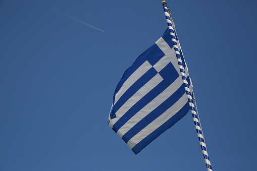 Greek flag on flagpole waving in the wind with clear blue sky behind Traditional colors of Greece: blue symbolizes the sky and the sea, white symbolizes the clouds and the waves.