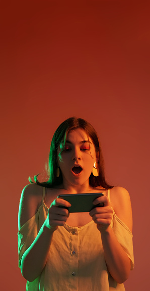 Smartphone playing. Amazed woman portrait. Surprised shocked neon color light girl playing online games isolated on red copy space background.