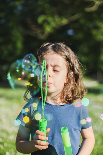 Portrait of a beautiful caucasian girl with brown short hair in a blue t-shirt blowing soap bubbles with colorful and magic bokeh while standing in a park on a playground with blurred trees background, close-up bottom view with elective focus. The concept of PARKS and REC, happy childhood, children's picnic, holidays, fabulous childhood, outdoor recreation, playgrounds, outdoors, soap bubbles.