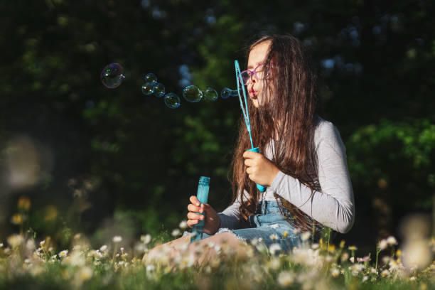 Portrait of a caucasian girl blowing soap bubbles while sitting in a flower meadow in a park Portrait of a beautiful caucasian girl blowing soap bubbles while sitting on a flower meadow in a park on a playground in the dark, close-up side view with selective focus and copy space. The concept of PARKS and REC, happy childhood, children's picnic, holidays, fabulous childhood, outdoor recreation, playgrounds, outdoors, soap bubbles, magic. funny camping signs pictures stock pictures, royalty-free photos & images