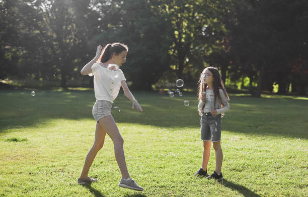 portrait of a beautiful caucasian girl sisters having fun, playing and blowing soap bubbles - outdoors playing family spring imagens e fotografias de stock