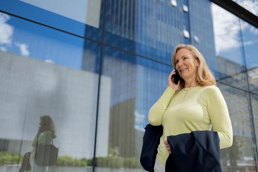 A casual business woman with a purse and a phone in her hand is walking in front of an office building, talking on the phone and in a good smiling mood. Sunny day, reflections of light in business glass buildings.