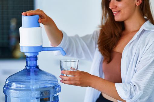 Portrait of young happy cute smiling woman pouring clean fresh water into glass from large bottle at home