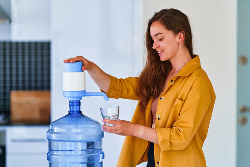Portrait of young happy cute smiling woman pouring clean fresh water into glass from large bottle at home