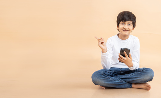 Happy Indian boy child holding smart phone in hand pointing finger at blank space to put advertisement, look here pose, standing over beige studio background,