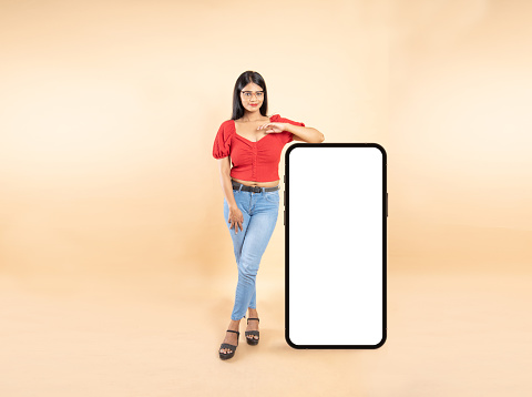 Beautiful Young Indian Woman Standing With Big Smart phone Blank White Screen Isolated On Beige Studio Background, Mobile Mockup, Copy Space.