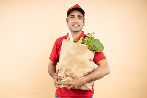 Indian delivery man wearing red uniform holding bag full of vegetables standing isolated over beige background. grocery delivery service concept.