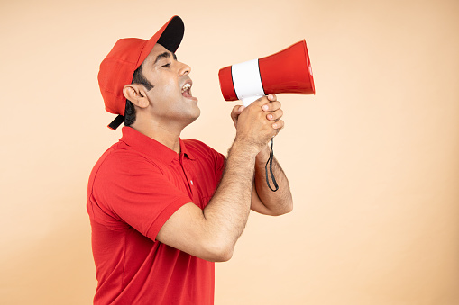 Happy excited Indian delivery man wearing red cap and T-shirt uniform hold screaming in megaphone or loud speaker isolated over beige Studio background, Shipping delivery and courier service concept.