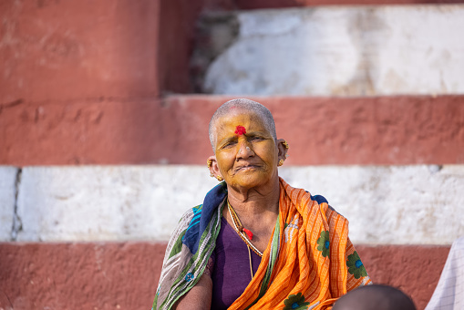 Varanasi, Uttar Pradesh, India - November 20 2022: Portrait on an old south indian woman with shaved head sitting on stairs at kedar ghat in traditional saree. Bald head is ritual performed in kashi.