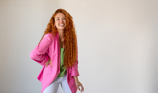 A stunning redhead latin girl with a beaming smile and freckled face stands against a white wall, wearing a pink blazer. Portrait of happy ginger young woman on white background with large copy space.
