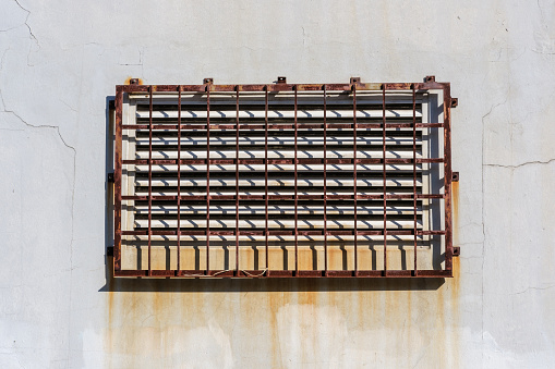 Rusty metal protective grate over a vented window in a white wall.