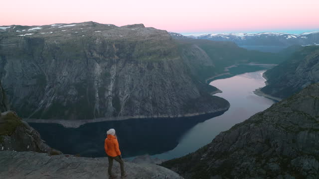 Aerial view: Woman hiker stand on top of rock overhanging above Fjord