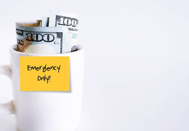 A white ceramic cup with cash dollars money inside, with orange paper sticky note written EMERGENCY ONLY, isolated on white background. Concept of savings for rainy day