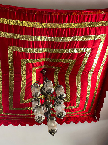 Stock photo showing close-up view of the ceiling of a domestic room with a glass and metal hurricane lampshade chandelier hanging under a gold and red fabric decoration.