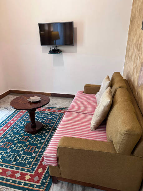 Stock photo showing a living room interior with a sofa covered with individual throw cushions and a wall hung smart screen television with floating shelf holding a VCR machine. Sofas are also commonly known as chesterfields, couches, futtons or settees. imagen de sofá tapizado beige con cojines de asiento a rayas rosas, cojines de tiro cuadrado, mesa de centro circular de madera sobre alfombra, televisión inteligente colgada en la pared, estante de acrílico transparente flotante que sostiene la máquin