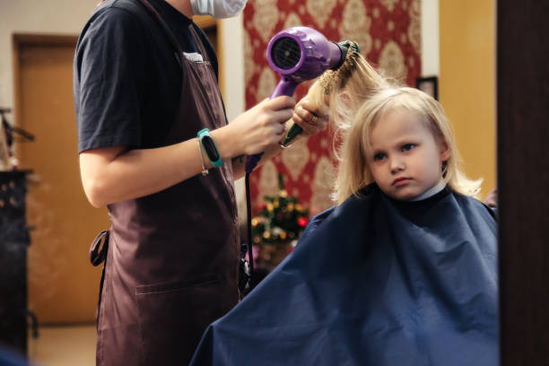Hair salon, hairdresser dries hair with hair dryer for kid in barber shop at mirror. Barber woman make hairstyle for cute little blonde girl in barbershop. Haircare, beauty concept. Copy ad text space peluquería, peluquería seca el cabello con secador de pelo para niños en peluquería en espejo. la mujer barbera hace peinado para la linda niña rubia en la barbería. cuidado del cabello, concepto de belleza. copiar el espacio de texto del anuncio