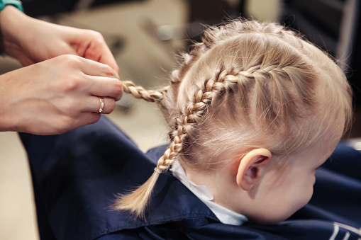 Close-up of hairdresser hands makes hairstyle for kid at barber shop. Hair salon, hairstylist braiding braids for pretty little blonde girl in barbershop. Haircare, beauty concept. Copy ad text space