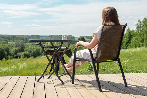Woman relaxing on terrace, sitting in chair at table, enjoying view on nature landscape, forest valley on summer holiday.