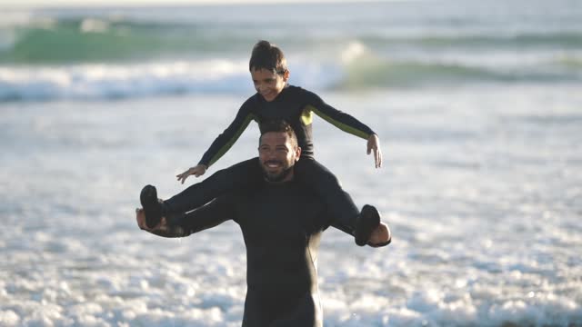 Smiling son on the shoulders of his father walking in the sea, playing airplanes and wearing wetsuits