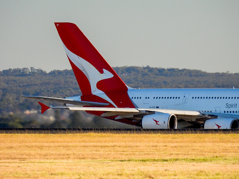 A Qantas Airbus A380-842 plane, registration VH-OQK, at the southern end of the main runway of Sydney Kingsford-Smith Airport and commencing her takeoff as flight number QF1 to Singapore.  This image was taken from Mill Stream Lookout on Botany Bay on a sunny, cold afternoon on 24 June 2023.